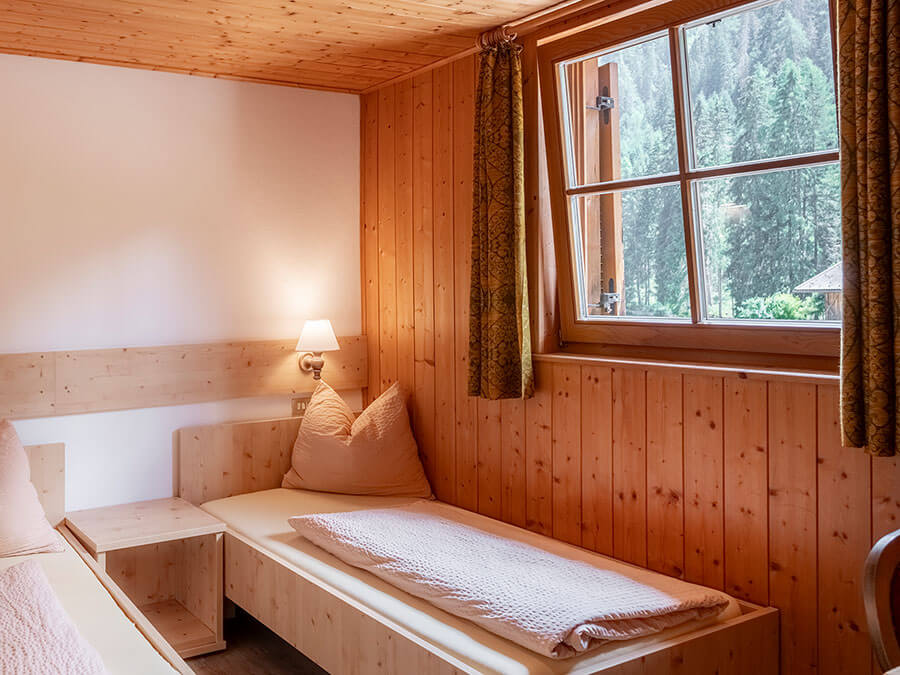Gemütliches Schlafzimmer aus Holz mit zwei Einzelbetten, einem Fenster und einer Lampe an der Wand zwischen den Betten. - Hotel Steinerhof