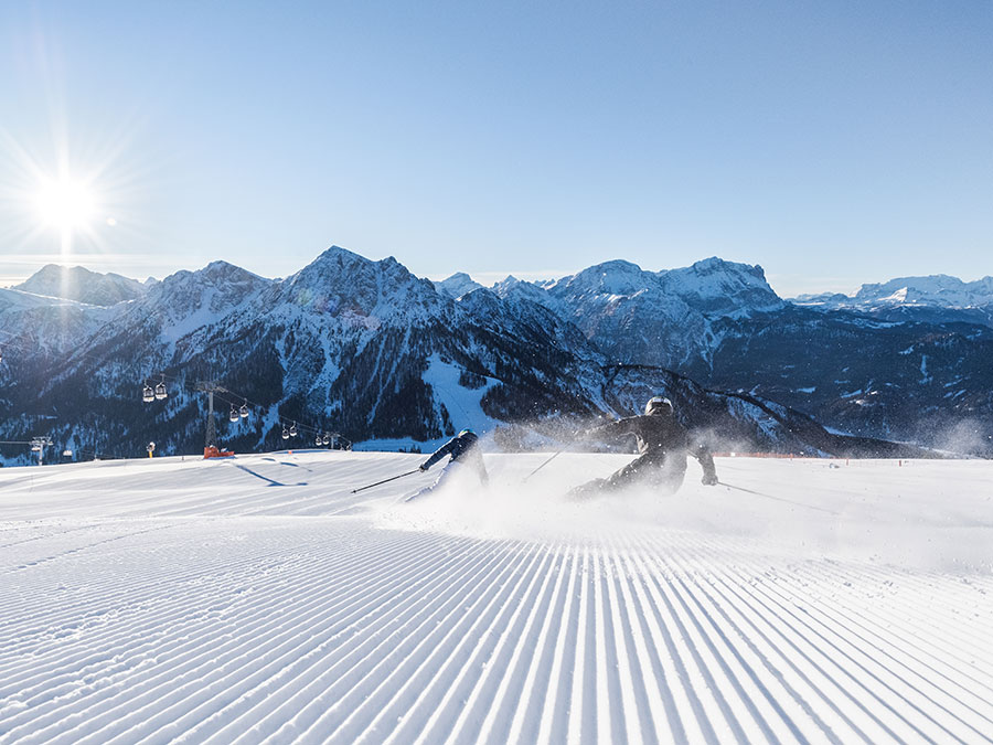 Ein Skifahrer fährt in Kurven auf einer präparierten Piste unter strahlendem Sonnenschein, im Hintergrund sind schneebedeckte Berge zu sehen. - Hotel Steinerhof