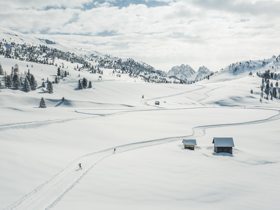Zwei Menschen fahren auf verschneiten Pisten in der Nähe kleiner Hütten in einer weiten, schneebedeckten Berglandschaft unter bewölktem Himmel Ski. - Hotel Steinerhof