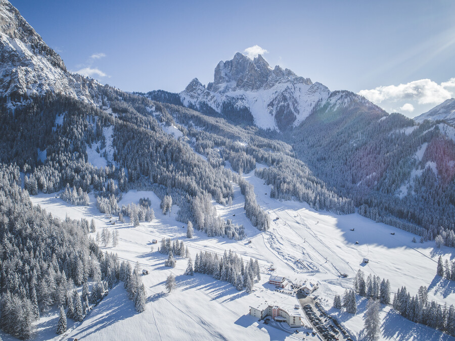 Vogelperspektive auf eine verschneite Berglandschaft mit einem kleinen Dorf und Kiefern unter strahlend blauem Himmel. - Hotel Steinerhof
