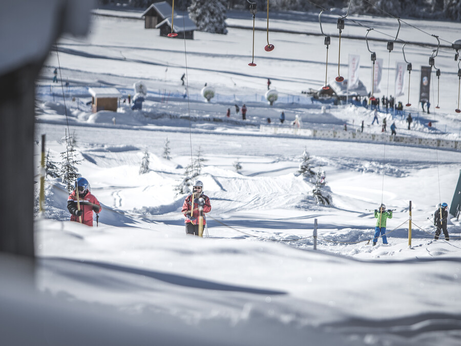 Skifahrer nutzen einen Knopflift auf einem verschneiten Hang, im Hintergrund sind Menschen und Gebäude zu sehen. - Hotel Steinerhof