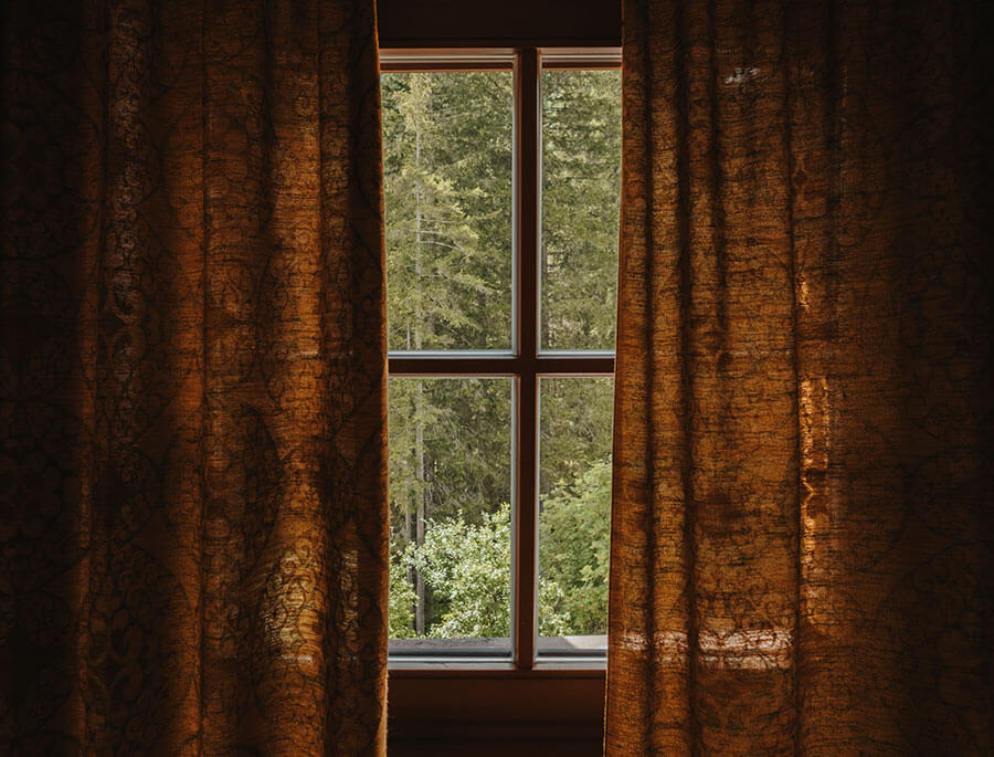 Braune Vorhänge rahmen ein Fenster ein, durch das man an einem sonnigen Tag den Blick auf grüne Bäume genießen kann. - Hotel Steinerhof