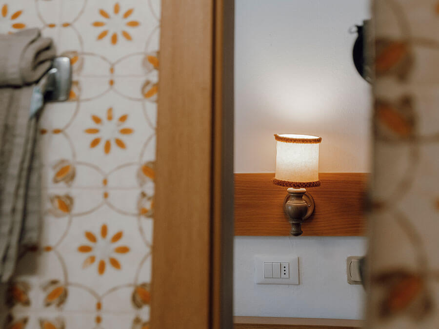 Eine Wandleuchte und ein Lichtschalter spiegeln sich in einem Spiegel wider; links sind eine gemusterte Tapete und ein Handtuch zu sehen. - Hotel Steinerhof