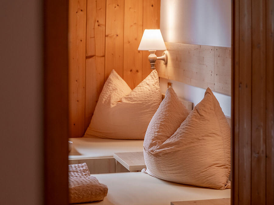 Gemütliches Schlafzimmer mit zwei Betten, beiger Bettwäsche, Holzwänden und einer eingeschalteten Wandlampe zwischen den Betten. - Hotel Steinerhof