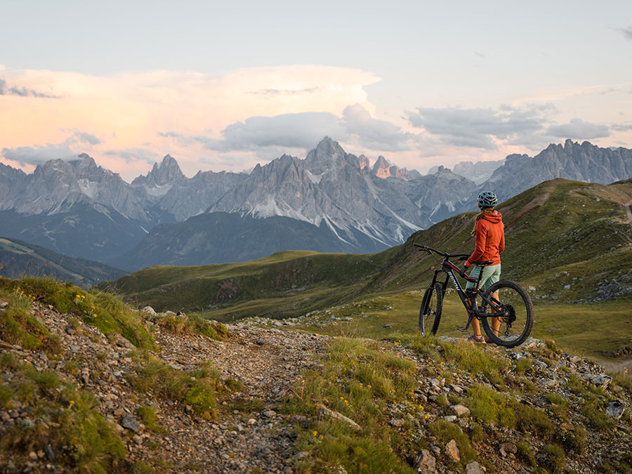 Eine Person mit einem Mountainbike steht auf einem felsigen Weg und blickt unter einem bewölkten Himmel auf die fernen Berge. - Hotel Steinerhof