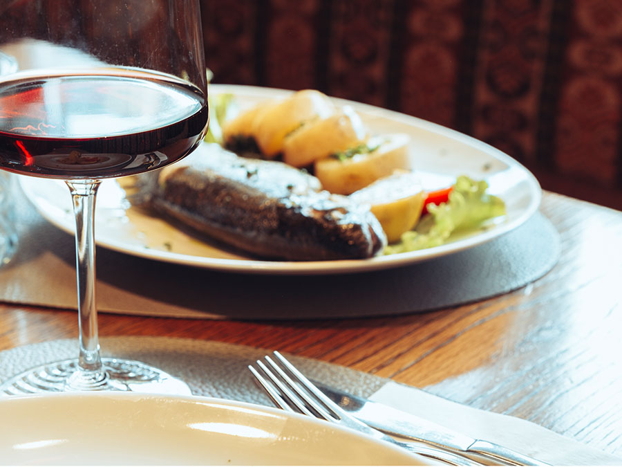Ein Glas Rotwein und ein Teller mit Fisch, Kartoffeln und Salat auf einem Holztisch in einem Restaurant. - Hotel Steinerhof