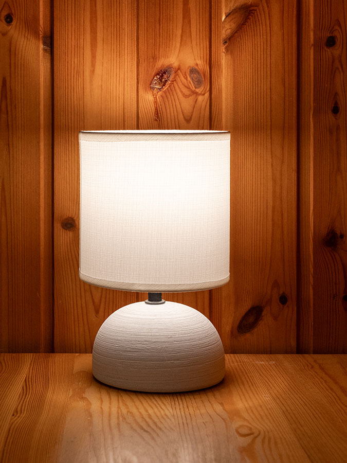 Auf einer Holzfläche vor einer holzvertäfelten Wand leuchtet eine weiße Tischlampe mit rundem Sockel. - Hotel Steinerhof