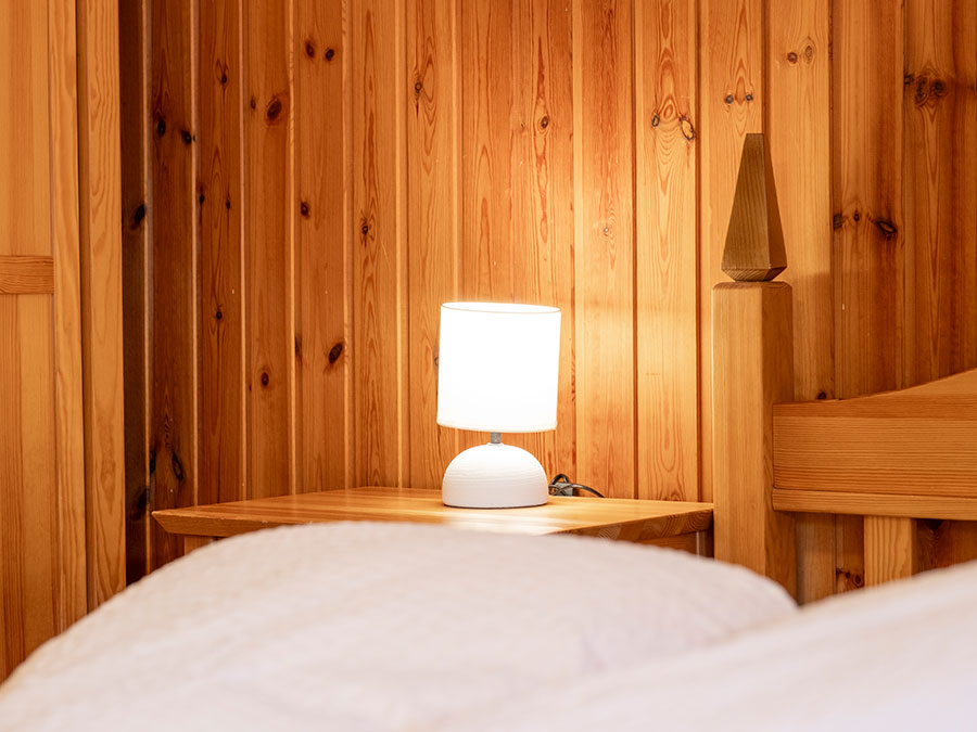 Eine weiße Lampe auf einem hölzernen Nachttisch neben einem Bett mit weißer Bettwäsche, vor einer hölzernen Wand. - Hotel Steinerhof
