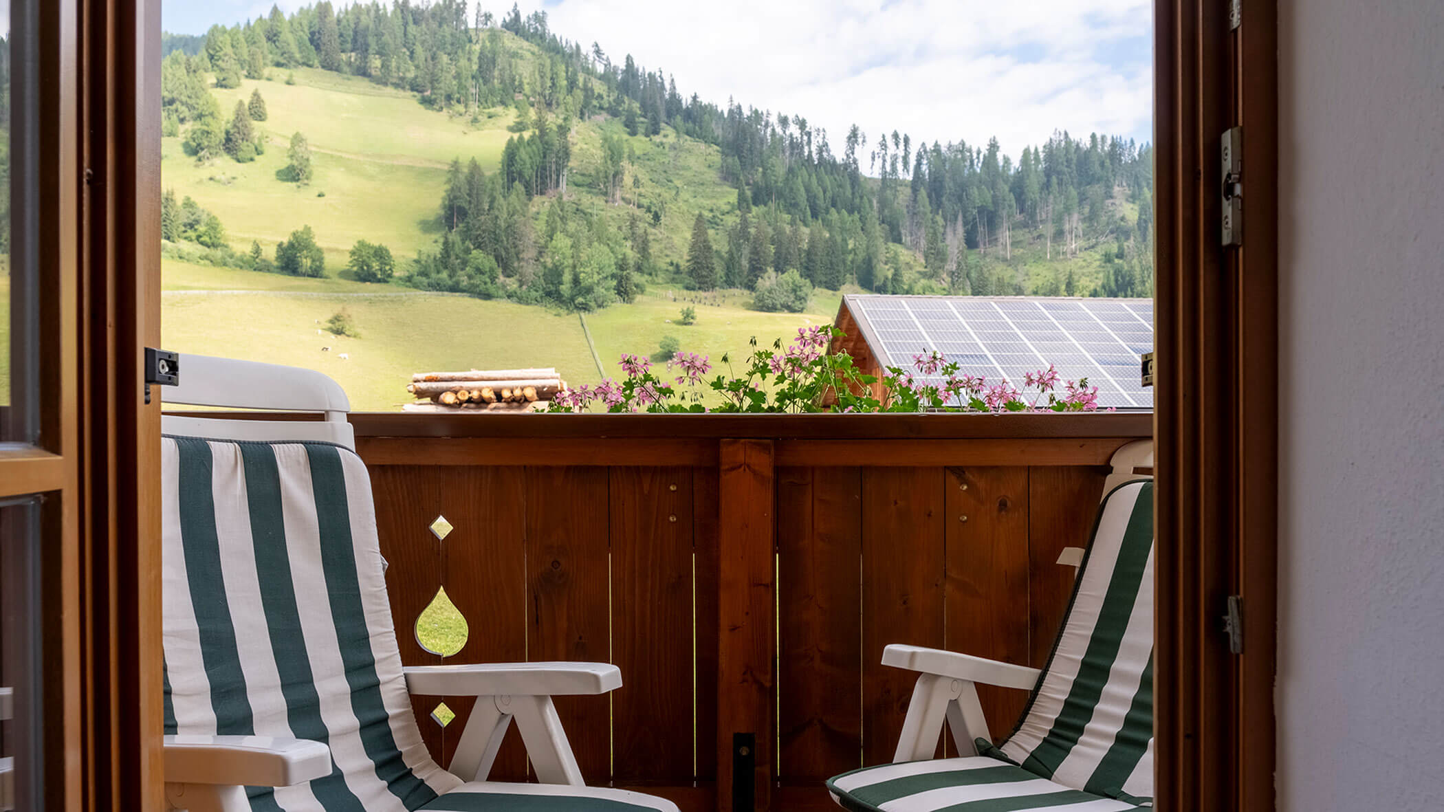 Von zwei gestreiften Stühlen auf einem Balkon aus blickt man auf grüne Hügel und Bäume, im Hintergrund ist ein Solardach zu sehen. - Hotel Steinerhof