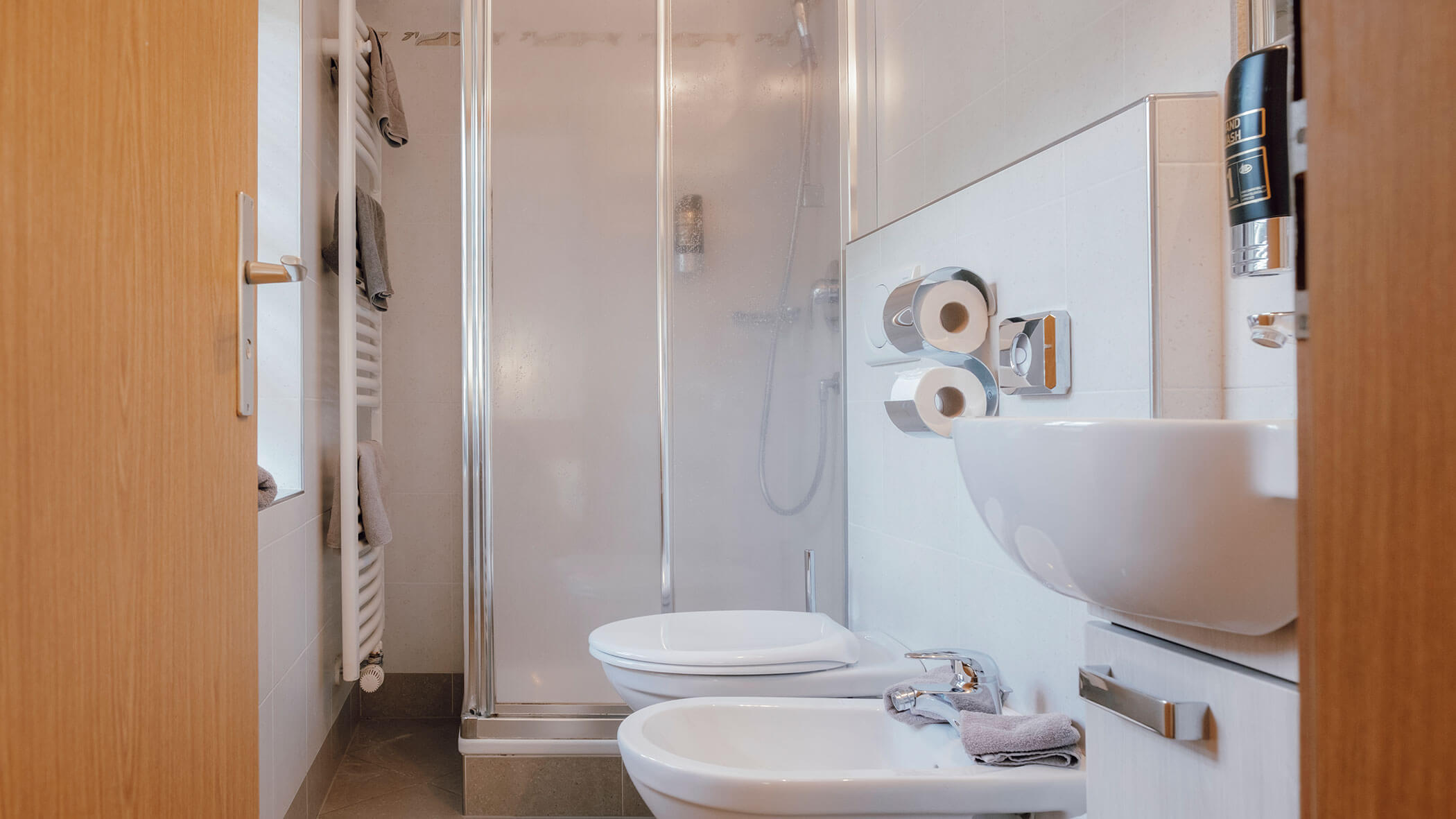 Kleines Badezimmer mit Dusche, Toilette, Bidet, Waschbecken und Handtüchern, die ordentlich auf Regalen und Haltern aufgereiht sind. - Hotel Steinerhof