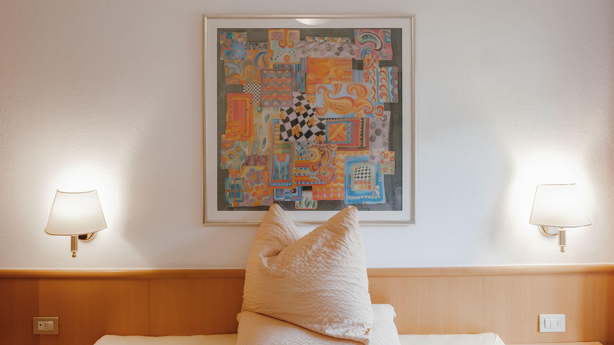 Ein Bett mit großen Kissen unter einem farbenfrohen abstrakten Gemälde, flankiert von zwei Wandleuchten. - Hotel Steinerhof