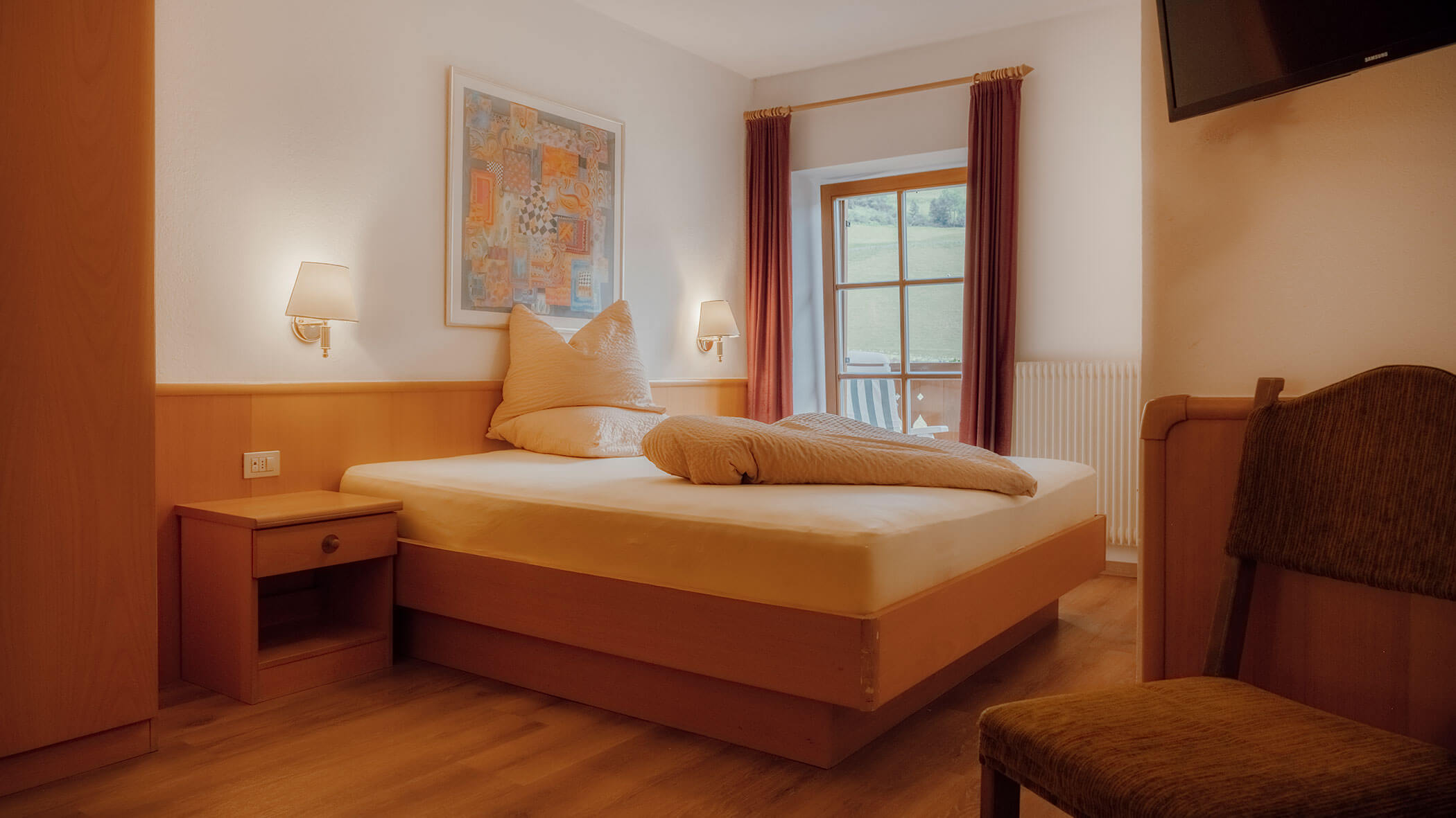 Gemütliches Hotelzimmer mit einem Doppelbett, einem Nachttisch, einem Fenster mit Vorhängen und einem Stuhl auf einem Holzboden. - Hotel Steinerhof