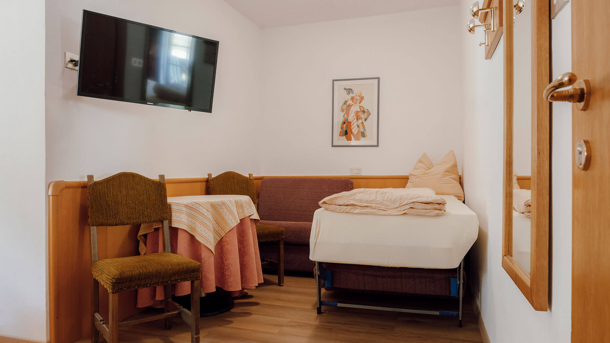 Kleines Zimmer mit einem Einzelbett, zwei Stühlen, einem Tisch, Wandbildern, einem Spiegel und einem an der Wand montierten Fernseher. - Hotel Steinerhof