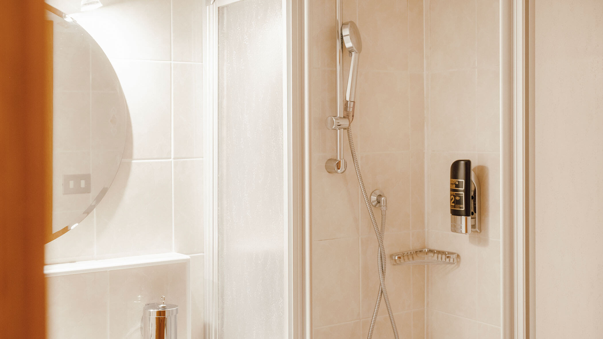 Eine mit beigen Fliesen ausgekleidete Dusche mit Handbrause, Seifenspender und einem runden Spiegel an der Wand. - Hotel Steinerhof