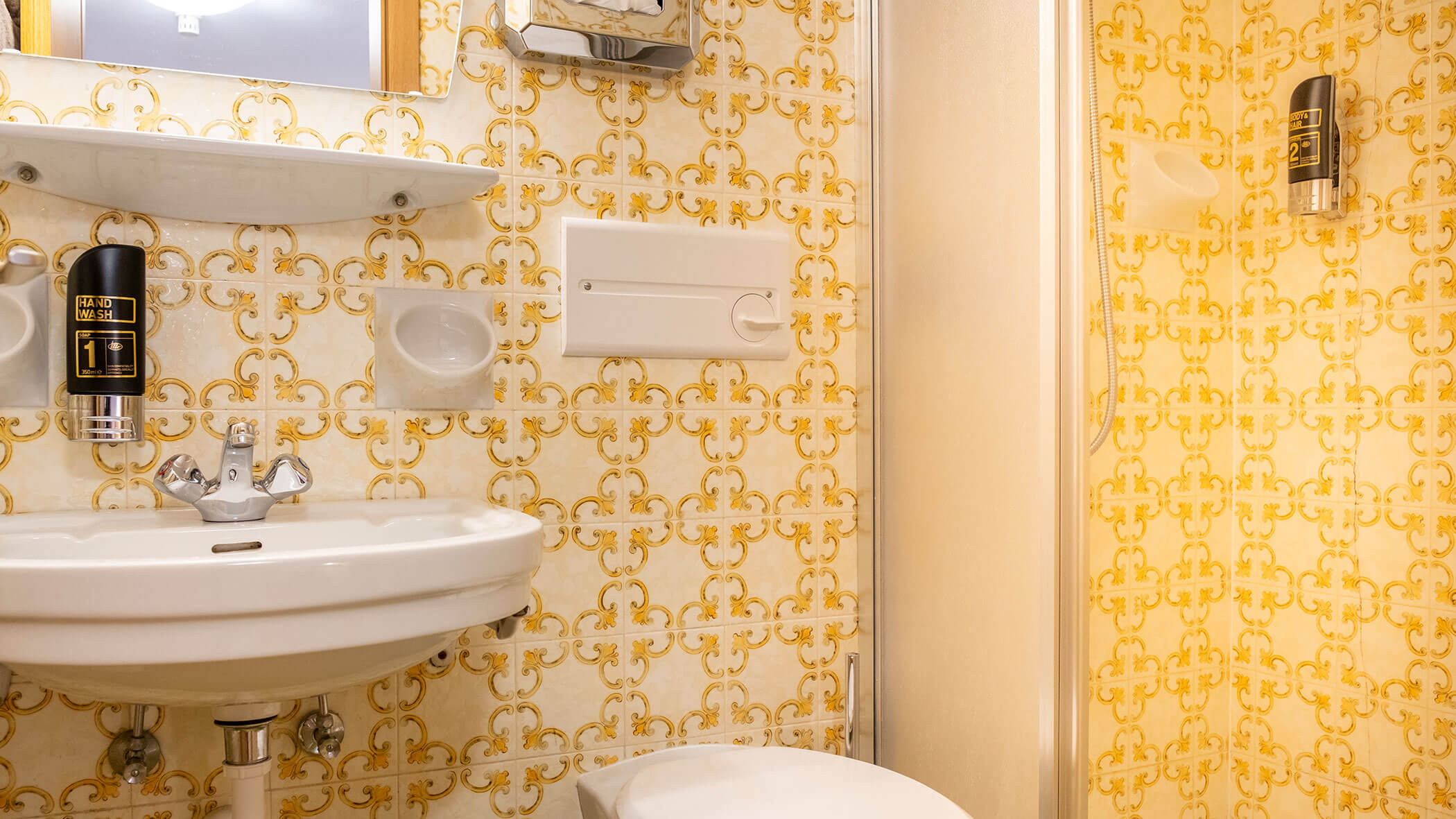 Kleines Badezimmer mit gelb gemusterten Fliesen, einem weißen Waschbecken, einem Spiegel, Seifenspendern und einer Toilette. - Hotel Steinerhof