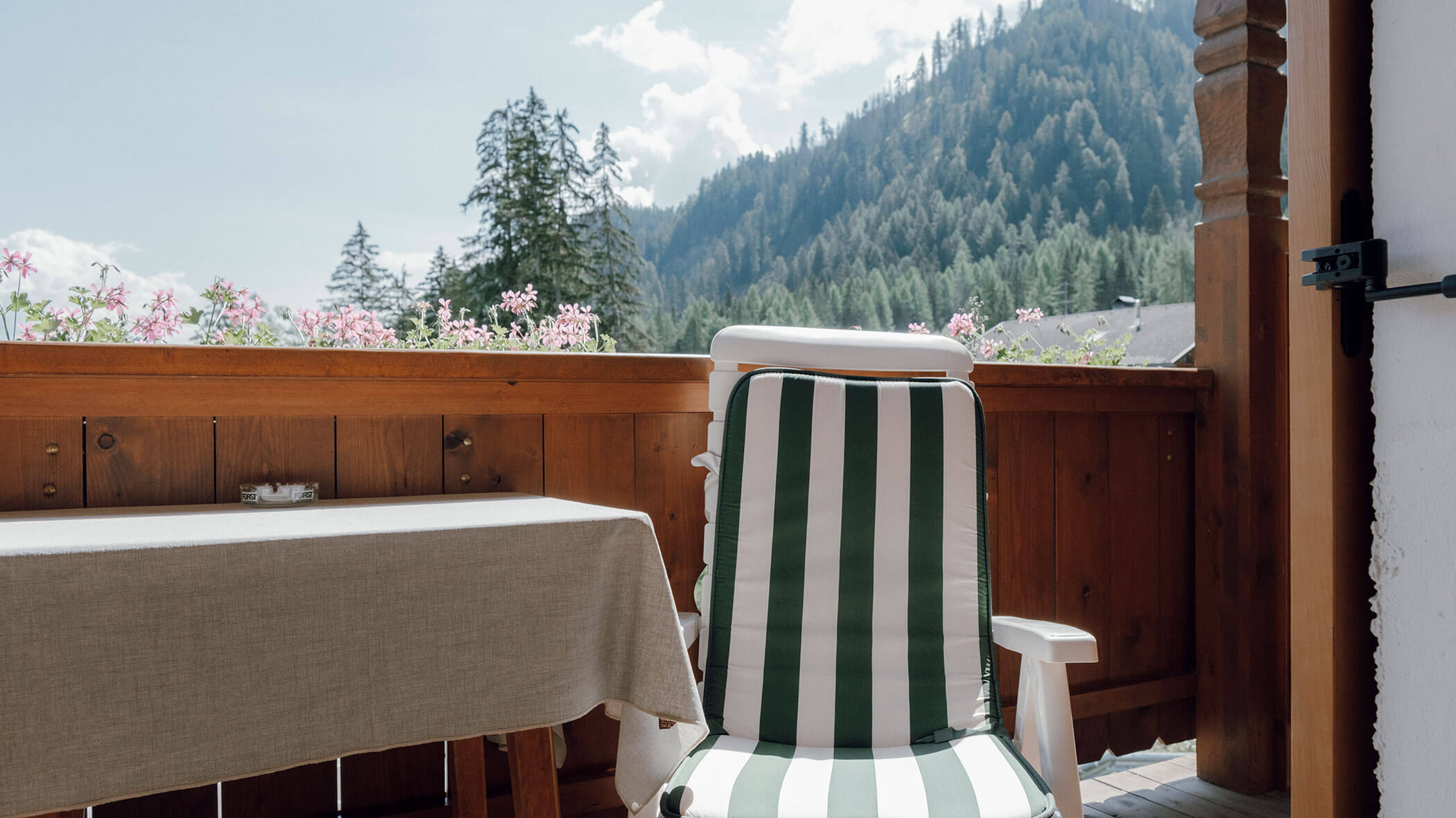 Ein gestreifter Stuhl und ein Tisch auf einem Holzbalkon bieten einen Blick auf mit Kiefern bewachsene Berge und den strahlend blauen Himmel. - Hotel Steinerhof