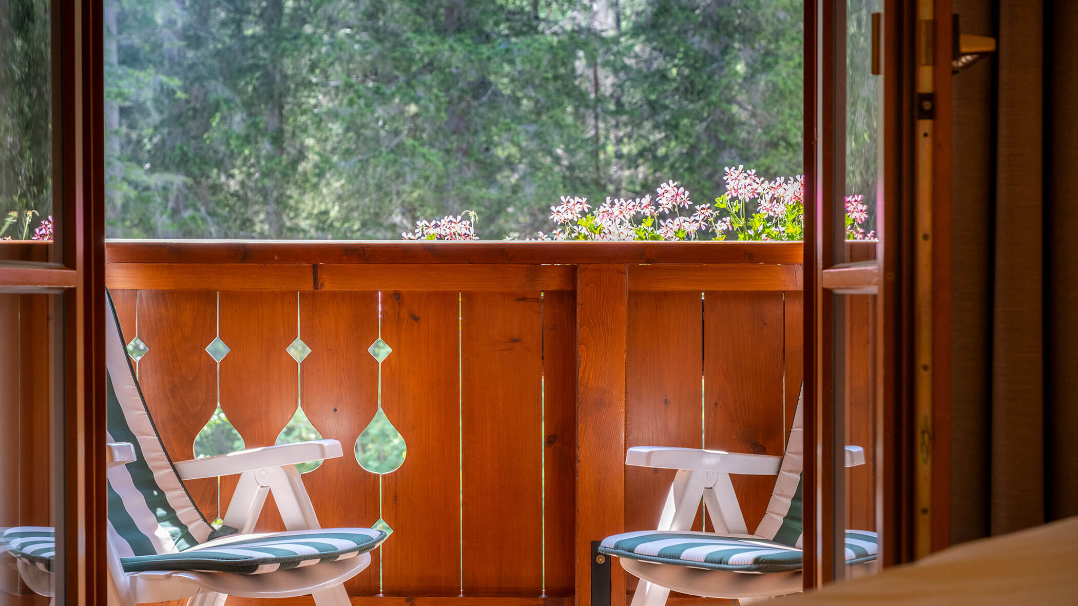 Zwei gestreifte Stühle auf einem hölzernen Balkon mit Blumen, im Hintergrund grüne Bäume. - Hotel Steinerhof