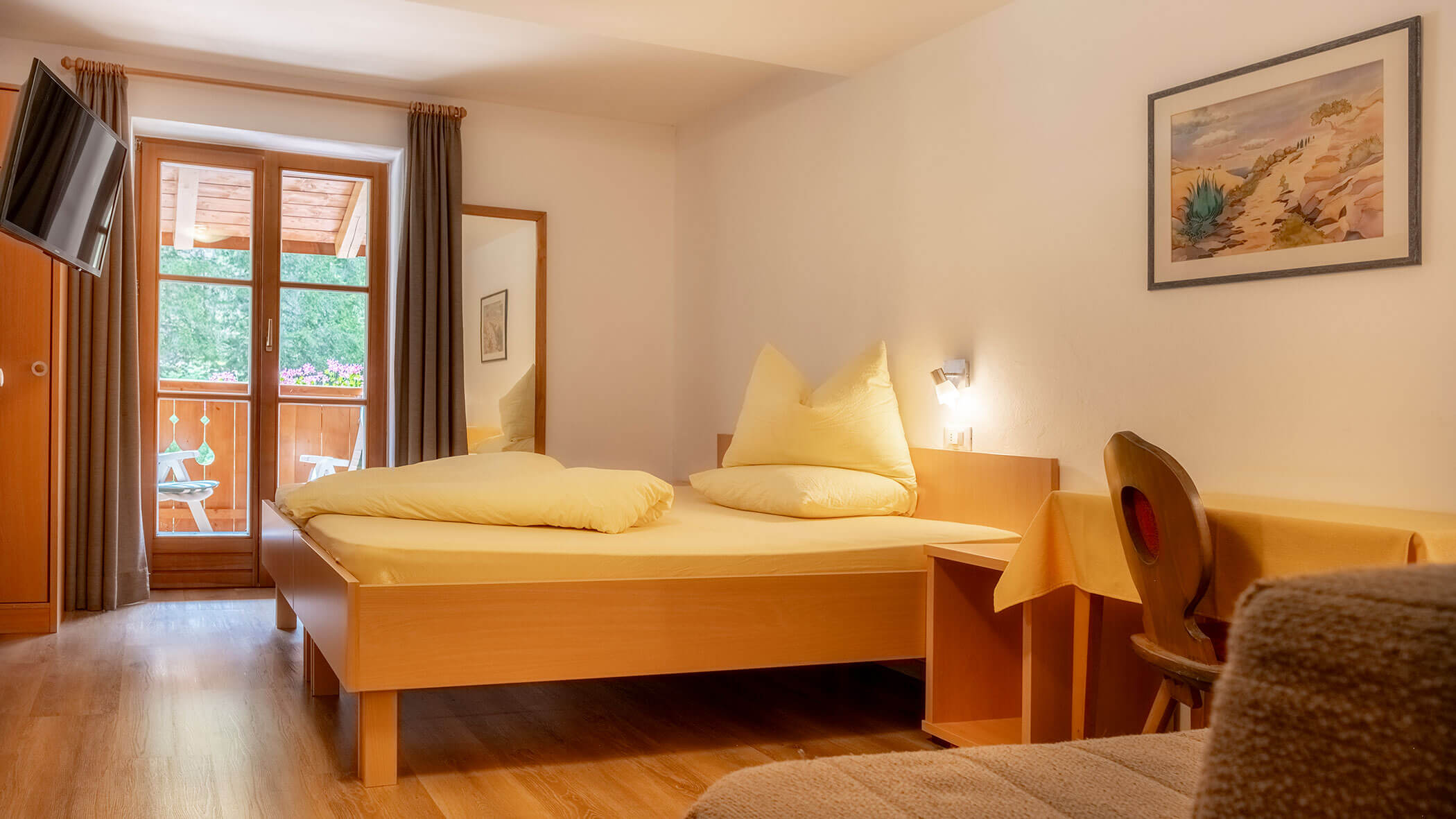 Gemütliches Schlafzimmer mit einem Doppelbett, Zugang zum Balkon, einem Schreibtisch, einem Fernseher und einem großen Spiegel am Fenster. - Hotel Steinerhof