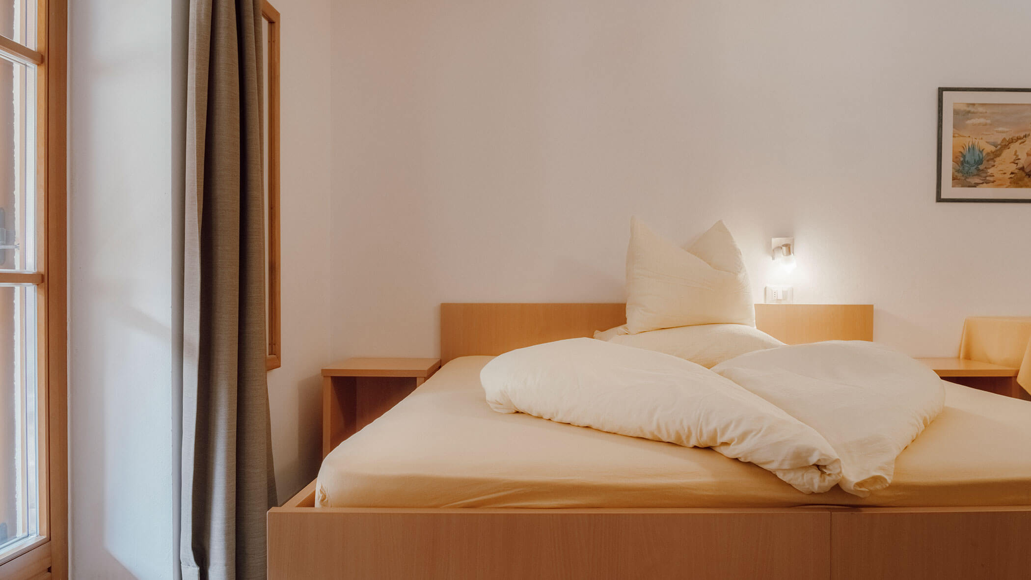 Ein ordentlich gemachtes Bett mit cremefarbener Bettwäsche in einem schlichten, hellen Schlafzimmer mit einem Fenster und Wandbildern. - Hotel Steinerhof