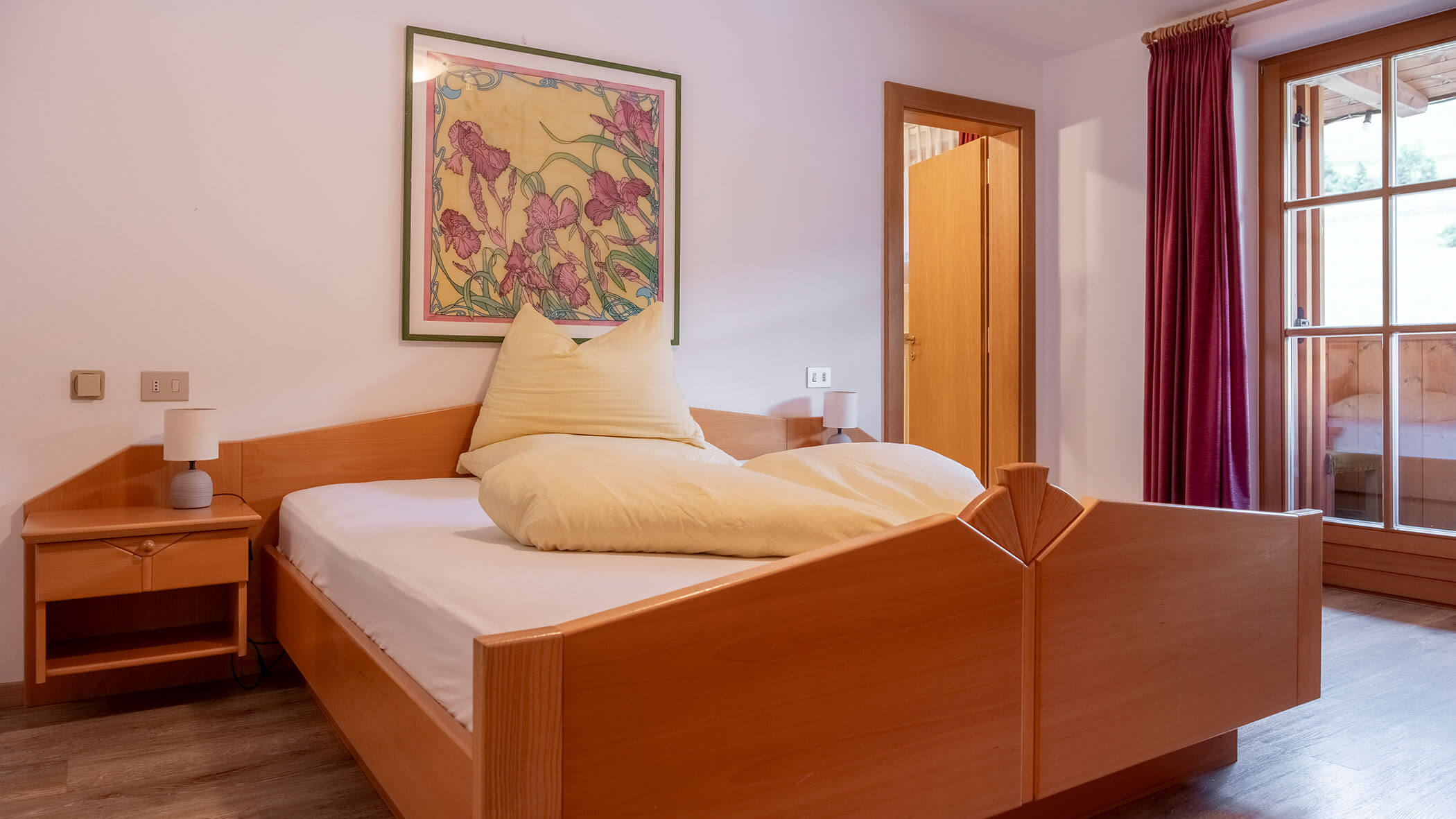 Ein Einzelbett mit gelber Bettwäsche, Holzmöbel, ein Nachttisch, Kunstwerke und ein Fenster mit roten Vorhängen. - Hotel Steinerhof