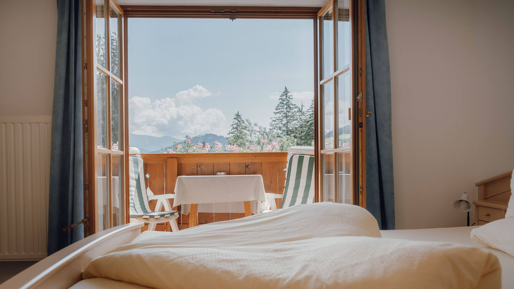Blick aus einem gemütlichen Schlafzimmer durch offene Türen auf einen Balkon mit Stühlen, einem Tisch, Blumen und Bergen. - Hotel Steinerhof