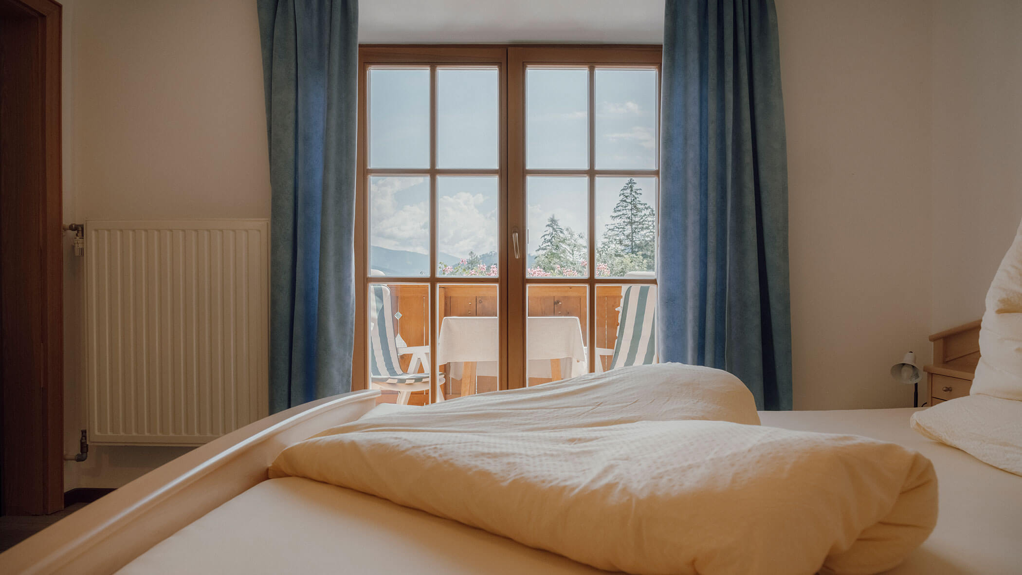 Ein ungemachtes Bett in einem Zimmer mit blauen Vorhängen, einem Balkon, Stühlen und Blick auf die Berge durch große Fenster. - Hotel Steinerhof