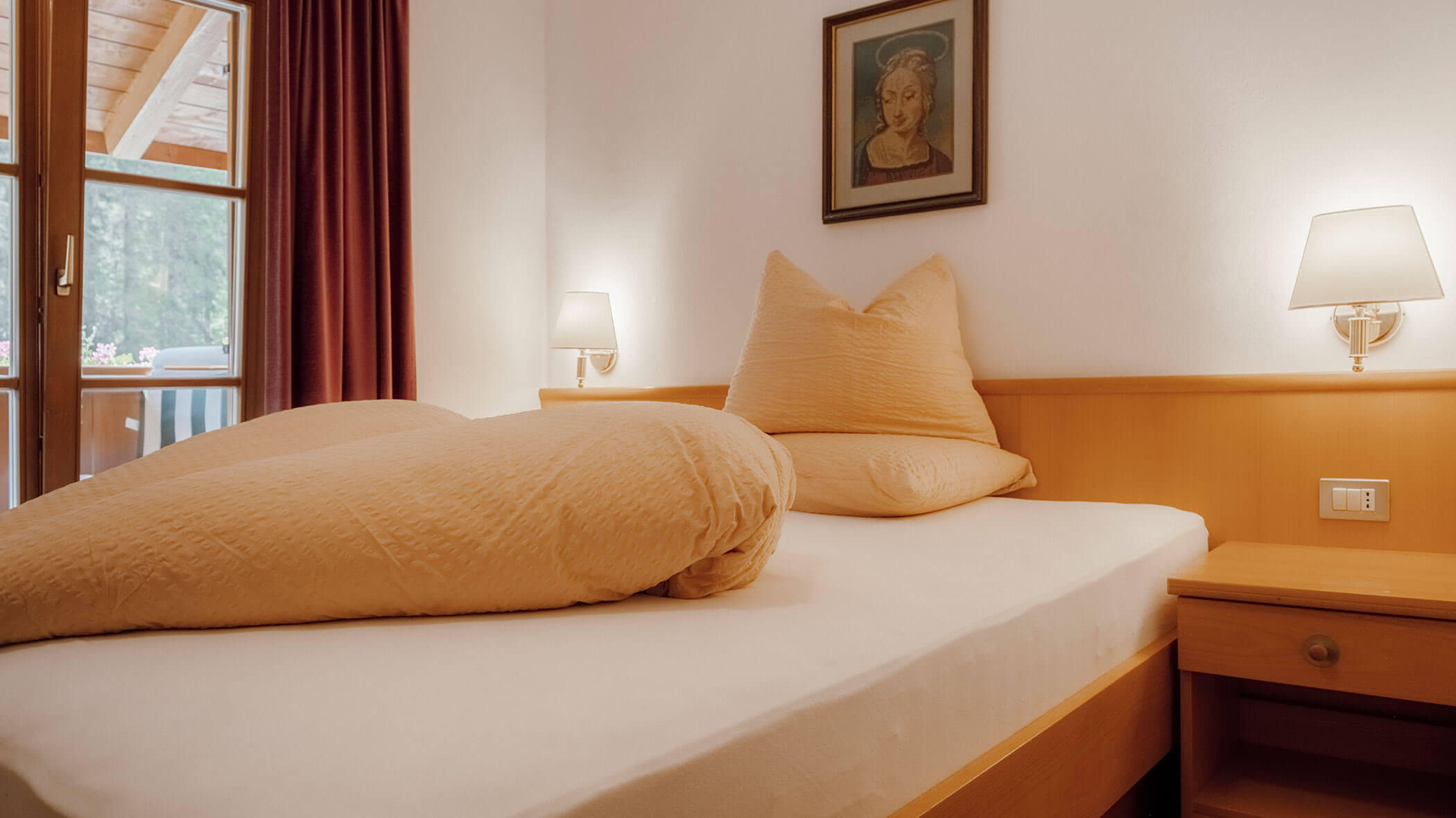 Gemütliches Schlafzimmer mit einem Doppelbett, pfirsichfarbener Bettwäsche, zwei Wandlampen, Kunstwerken und einem Fenster mit Vorhängen. - Hotel Steinerhof