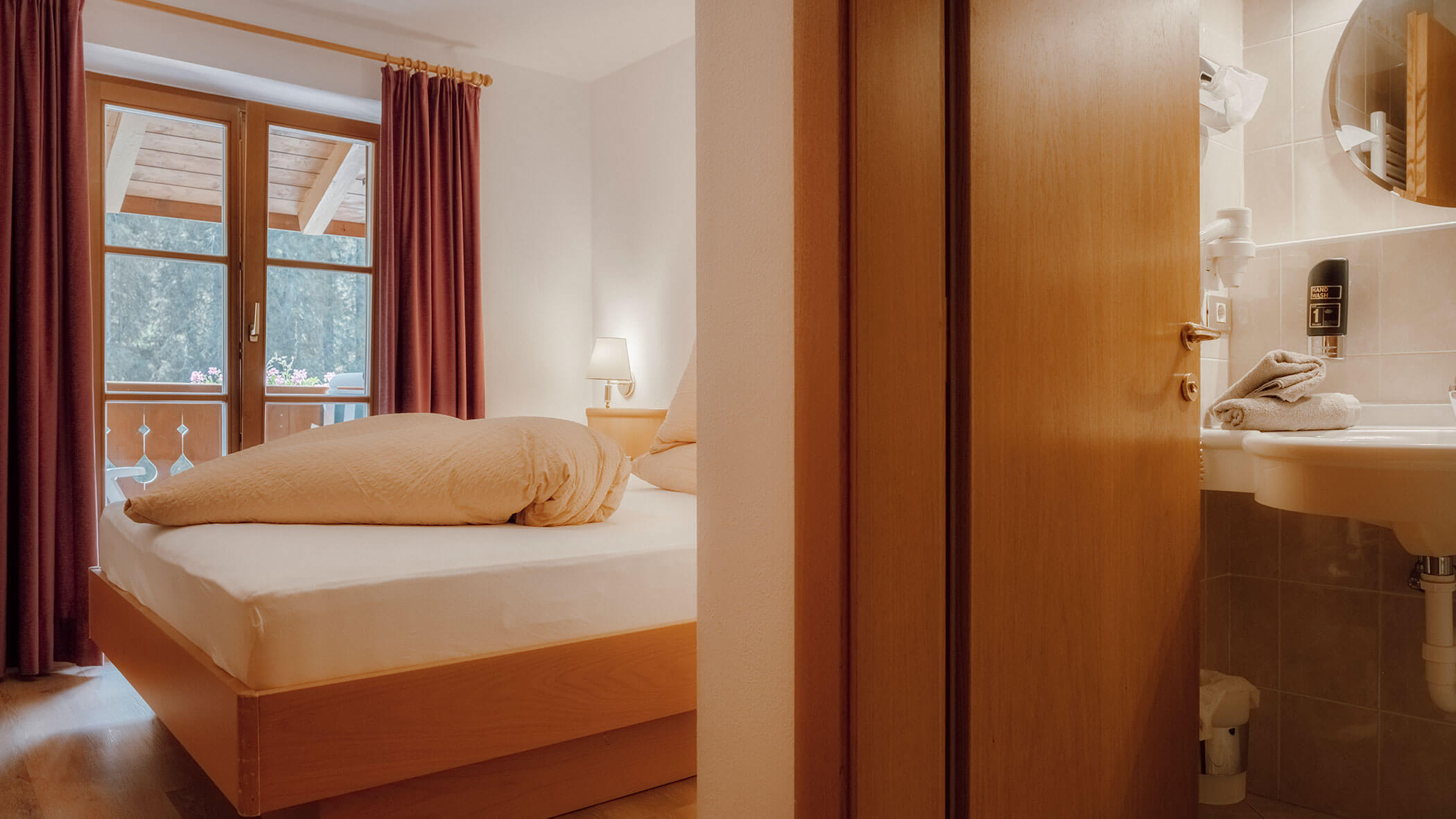 Gemütliches Schlafzimmer mit einem gemachten Bett, einem Fenster mit roten Vorhängen und Blick ins Badezimmer mit einem Waschbecken. - Hotel Steinerhof