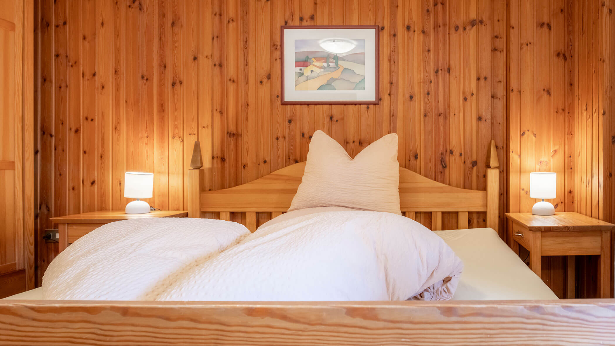 Gemütliches Schlafzimmer aus Holz mit einem Doppelbett, weißer Bettwäsche, zwei Nachttischen und Lampen sowie einem Gemälde an der Wand. - Hotel Steinerhof
