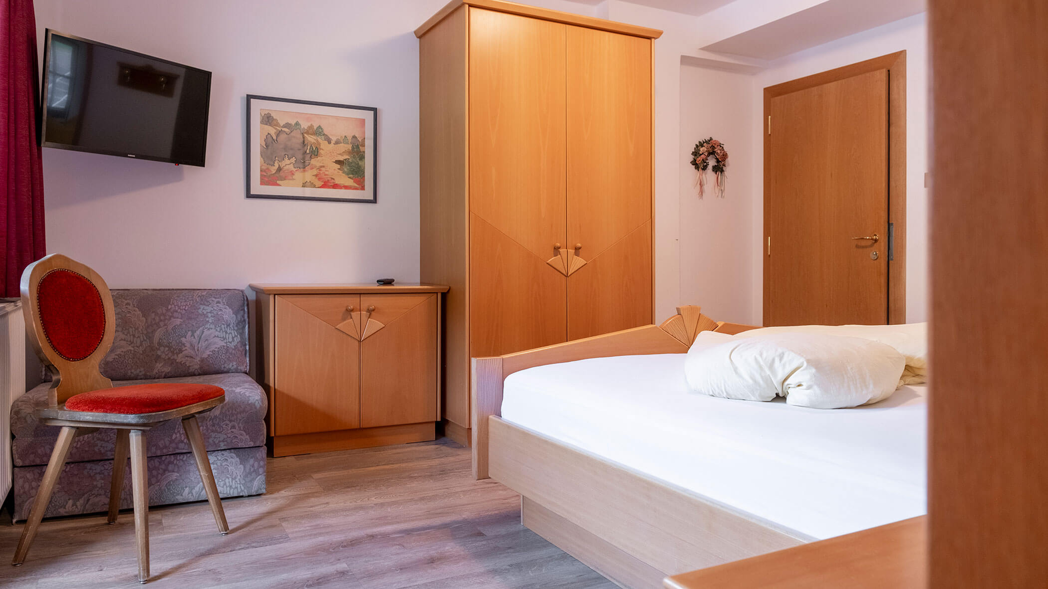 Ein gepflegtes Hotelzimmer mit einem Bett, einem Kleiderschrank, einem Fernseher, einem Sessel, einem kleinen Sofa und Holzmöbeln. - Hotel Steinerhof