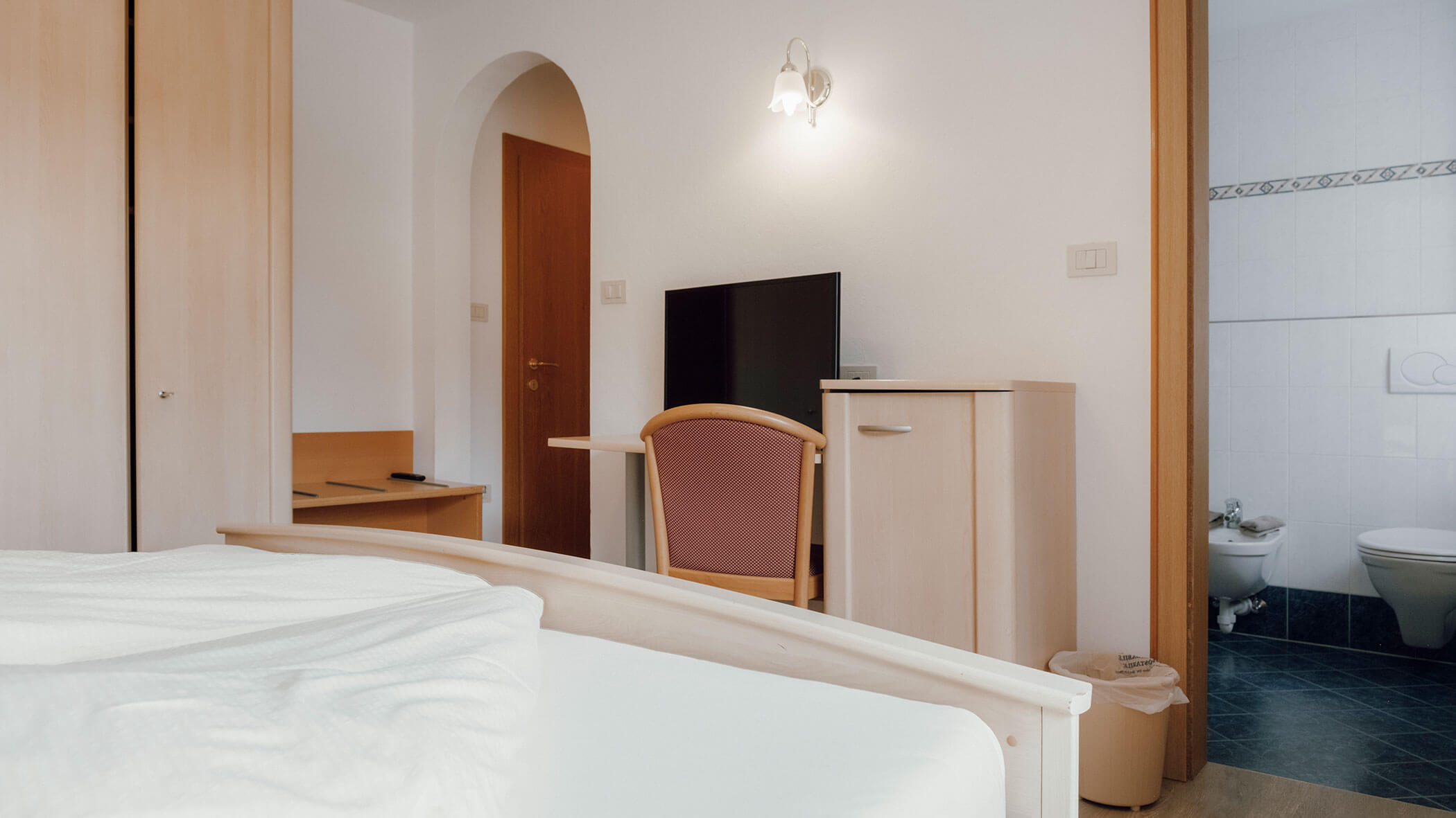Ein aufgeräumtes Hotelzimmer mit einem Bett, einem Schreibtisch, einem Stuhl, einem Fernseher, einem Schrank und einem Badezimmer, das im Hintergrund zu sehen ist. - Hotel Steinerhof