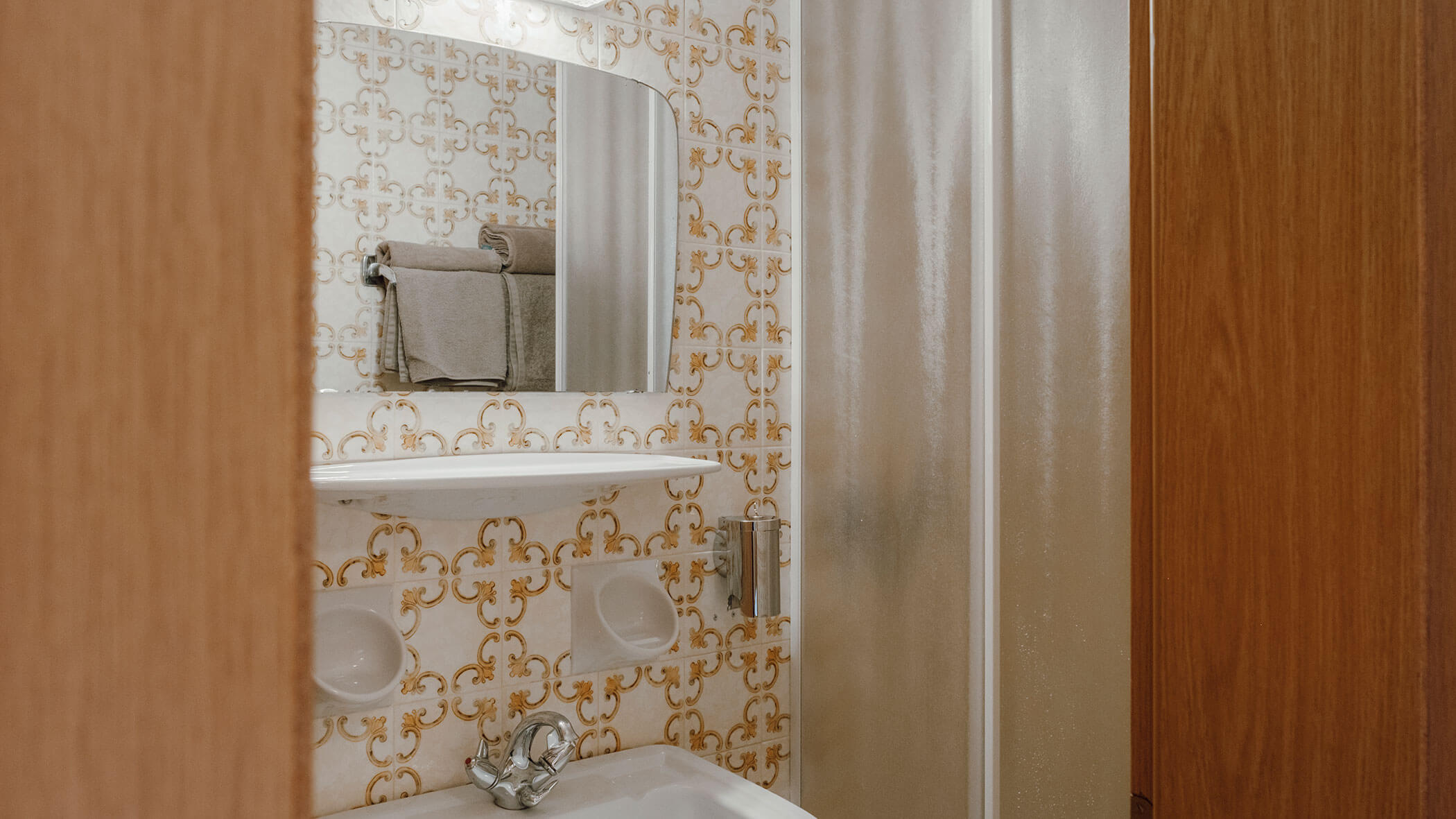 Kleines Badezimmer mit Fliesen im Vintage-Stil, einem Spiegel, einem Waschbecken, einem Regal, Handtüchern und einer mattierten Duschkabinentür. - Hotel Steinerhof