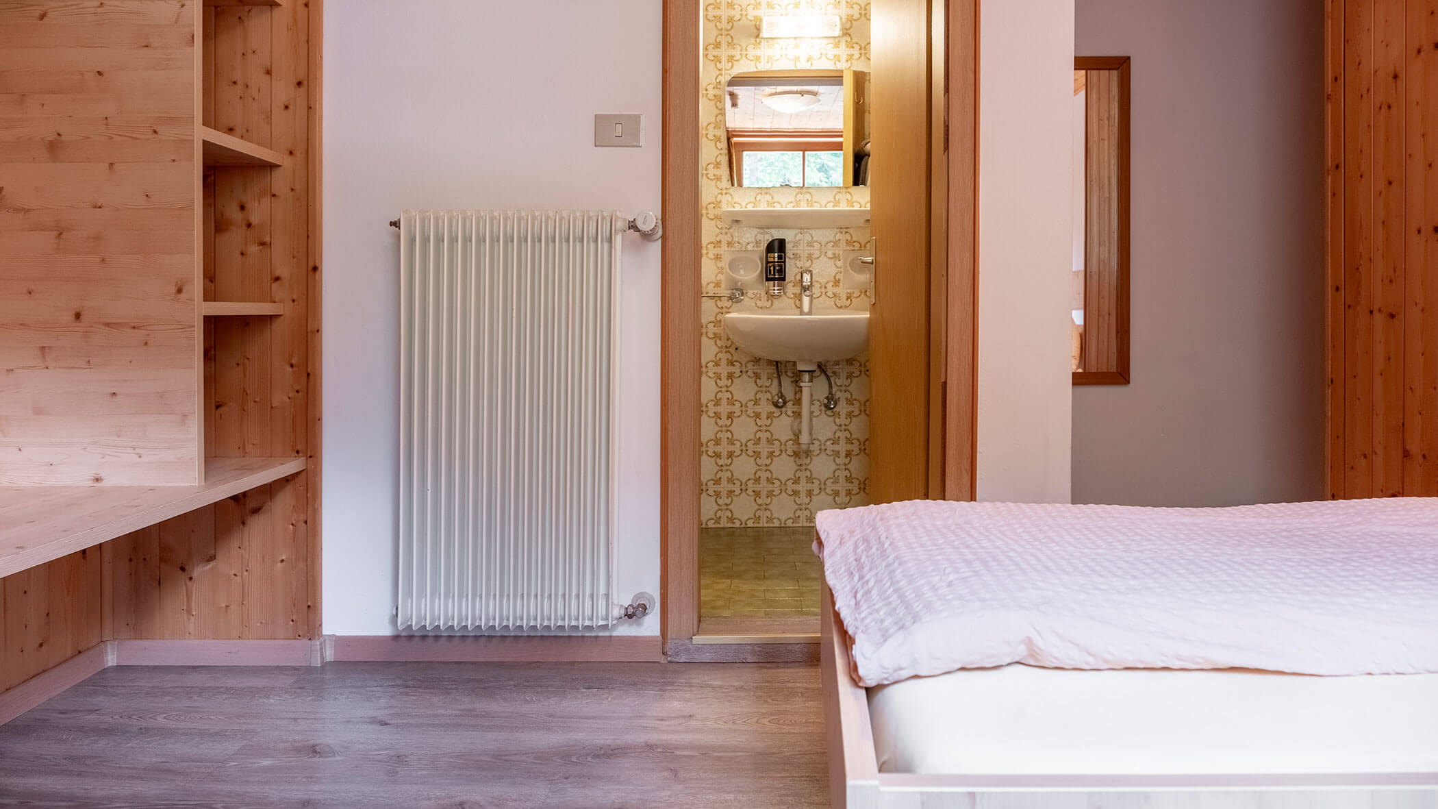 Ein Schlafzimmer mit einem Bett, einem Heizkörper und einer offenen Tür, durch die man ein Badezimmer mit einem Waschbecken und gefliesten Wänden sehen kann. - Hotel Steinerhof