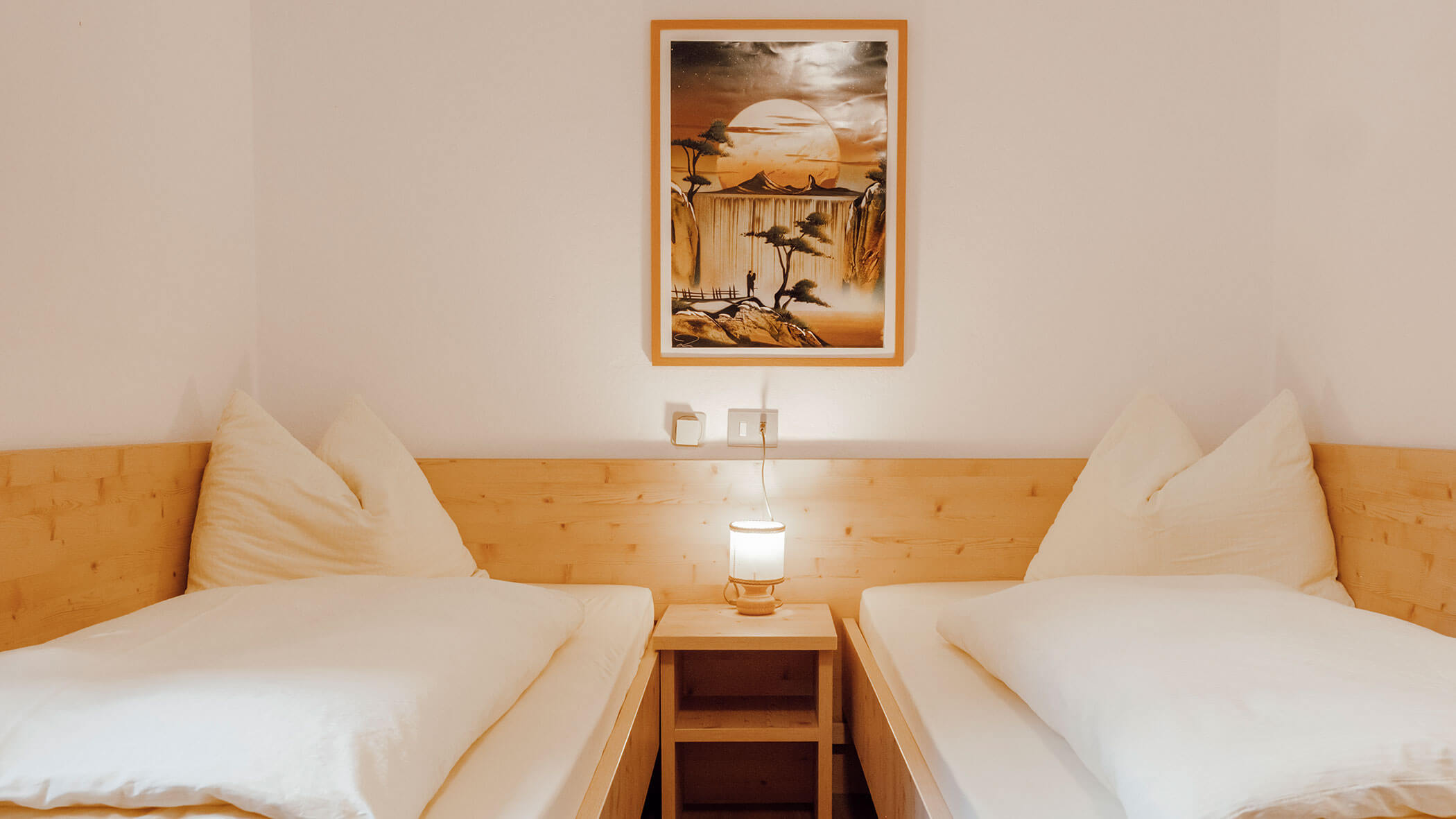 Zwei Einzelbetten mit weißer Bettwäsche, eine Lampe auf einem Nachttisch und ein gerahmtes Bild eines Sonnenuntergangs an der Wand. - Hotel Steinerhof
