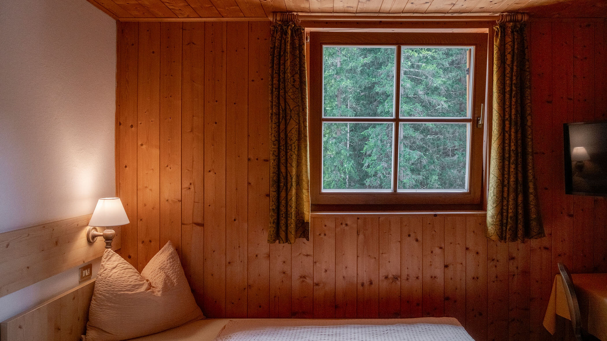 Gemütliches Holzzimmer mit einem Bett, einer Lampe, einem Fenster mit Blick auf grüne Bäume und teilweise geöffneten Vorhängen. - Hotel Steinerhof