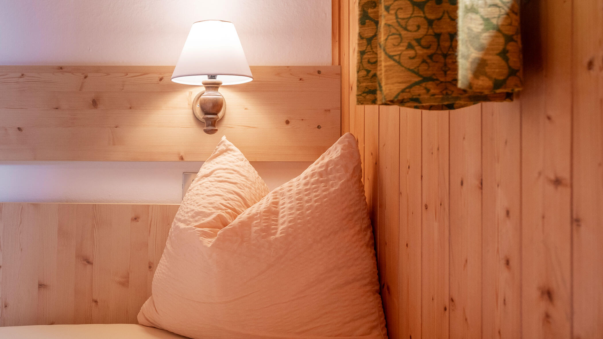 Ein gemütliches Bett mit einem großen Kissen, holzvertäfelte Wände, eine Wandlampe und ein grün gemusterter Vorhang. - Hotel Steinerhof