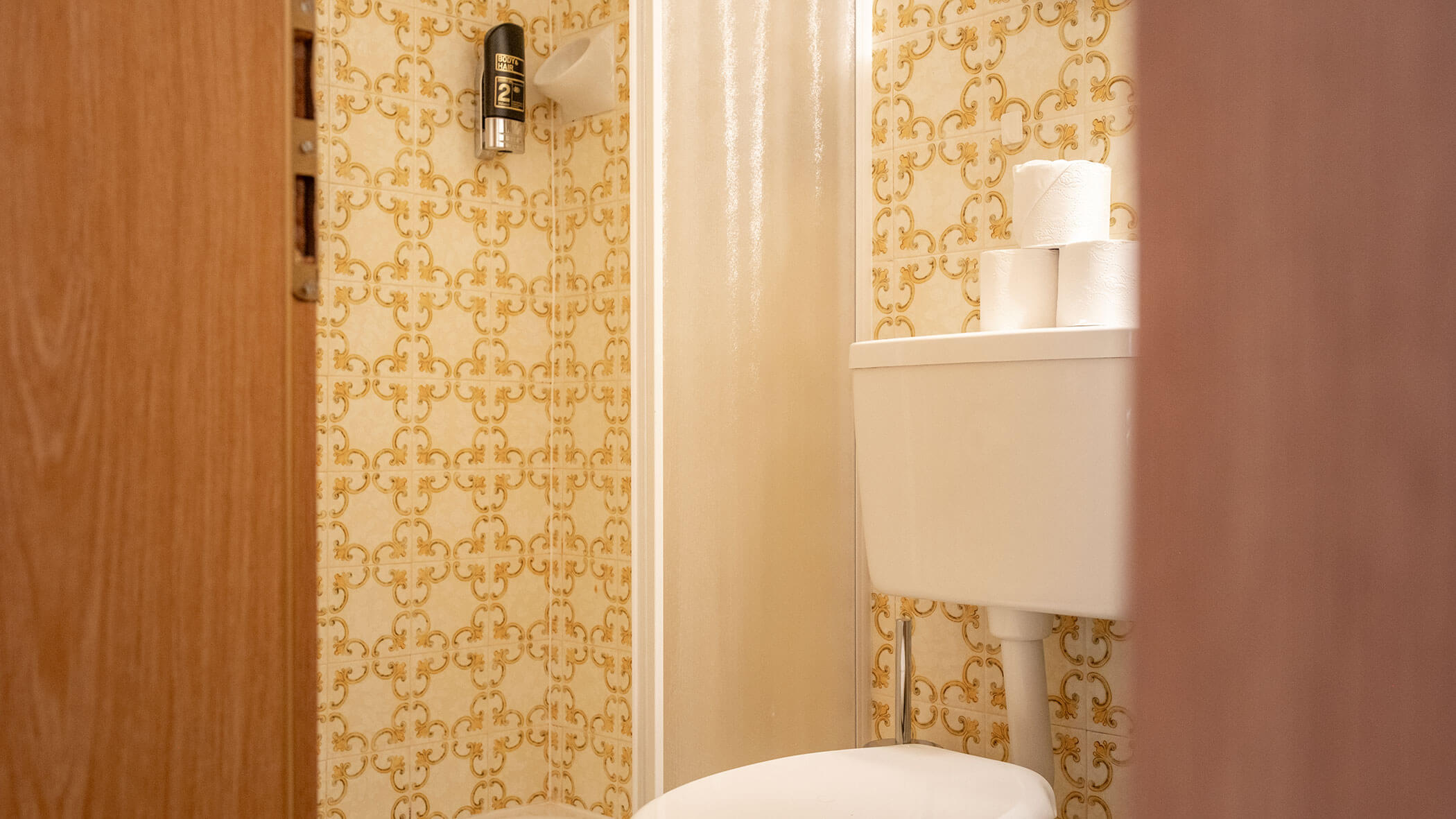 Kleines Badezimmer mit einer gelben Retro-Tapete mit Muster, einer weißen Toilette und gestapelten Toilettenpapierrollen. - Hotel Steinerhof