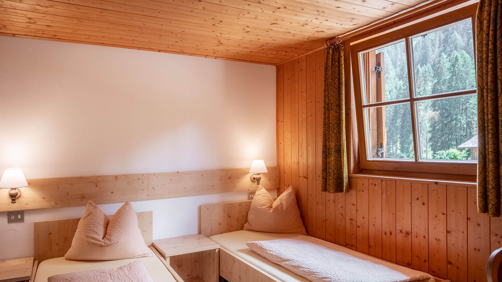 Gemütliches Zimmer mit zwei Einzelbetten, Holzwänden, Wandlampen und einem Fenster mit braunen Vorhängen, von dem aus man auf Bäume blickt. - Hotel Steinerhof