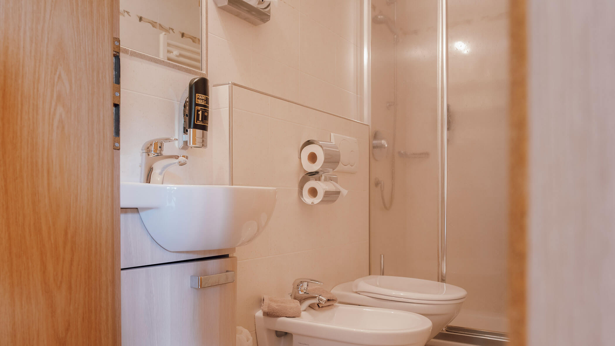 Kleines Badezimmer mit Waschbecken, Bidet, Toilette, Dusche, Toilettenpapier und einem an der Wand montierten Seifenspender. - Hotel Steinerhof