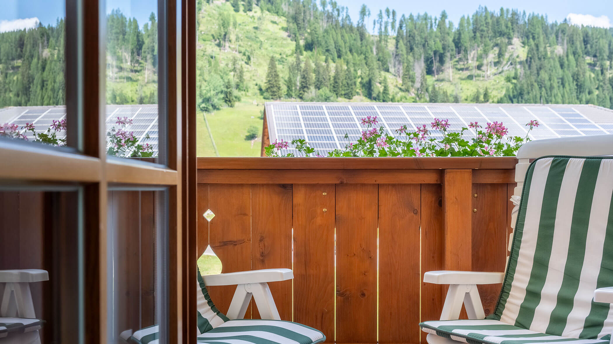 Balkon mit gestreiften Stühlen, Holzgeländer, Blumen und Solarmodulen, im Hintergrund Bäume. - Hotel Steinerhof