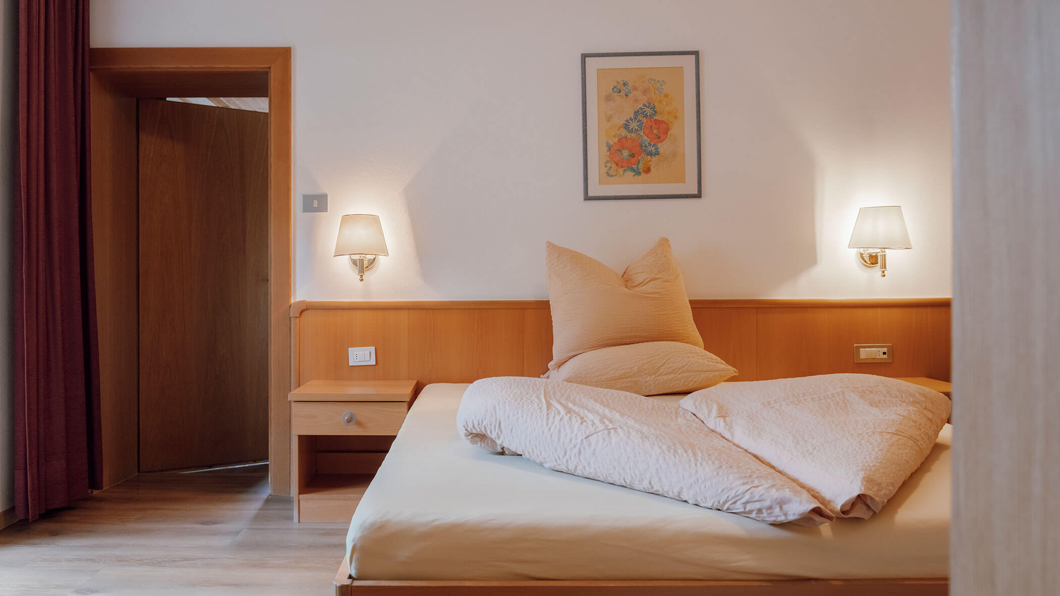Ein ordentlich gemachtes Bett mit beiger Bettwäsche, zwei Wandlampen und einem Blumenbild über dem Kopfteil. - Hotel Steinerhof