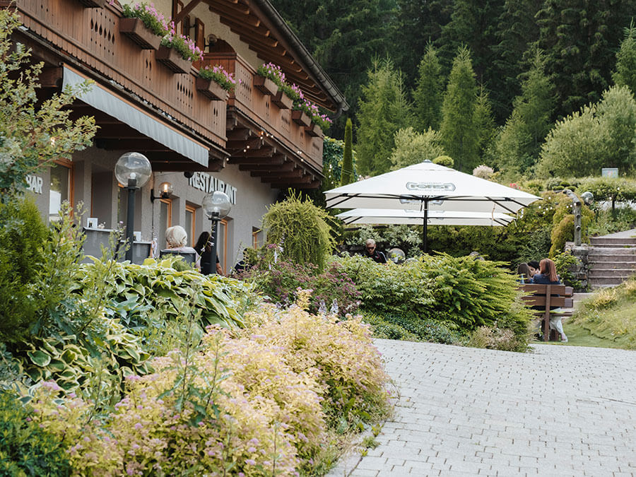 Menschen, die vor einem Restaurant mit Pflanzen, einem Sonnenschirm und einem bewaldeten Hintergrund im Freien essen. - Hotel Steinerhof