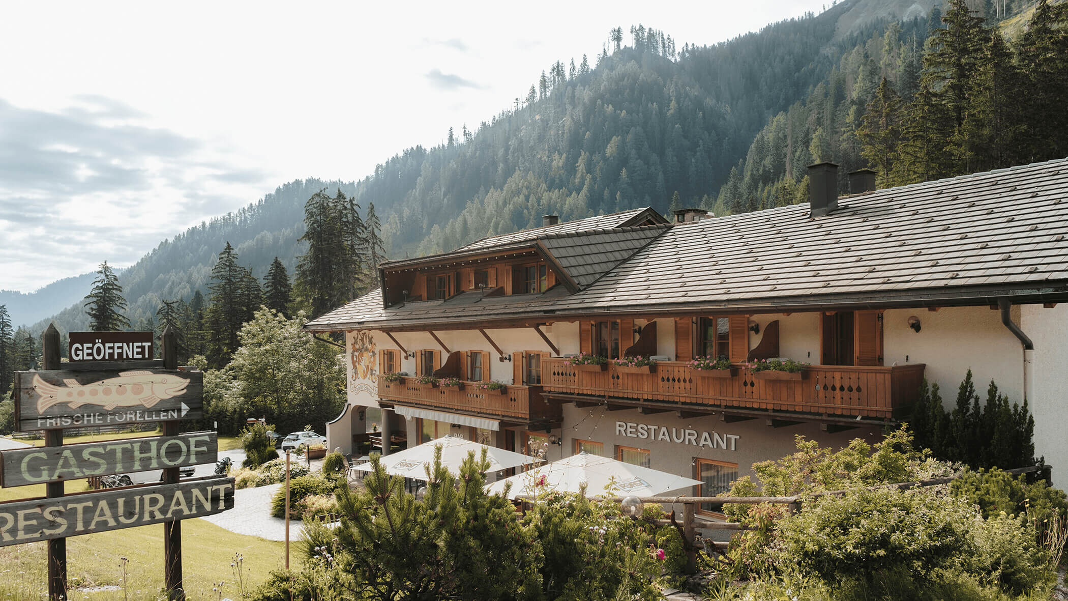 Alpenrestaurant mit Holzbalkonen, umgeben von Bäumen und Bergen unter strahlend blauem Himmel. - Hotel Steinerhof