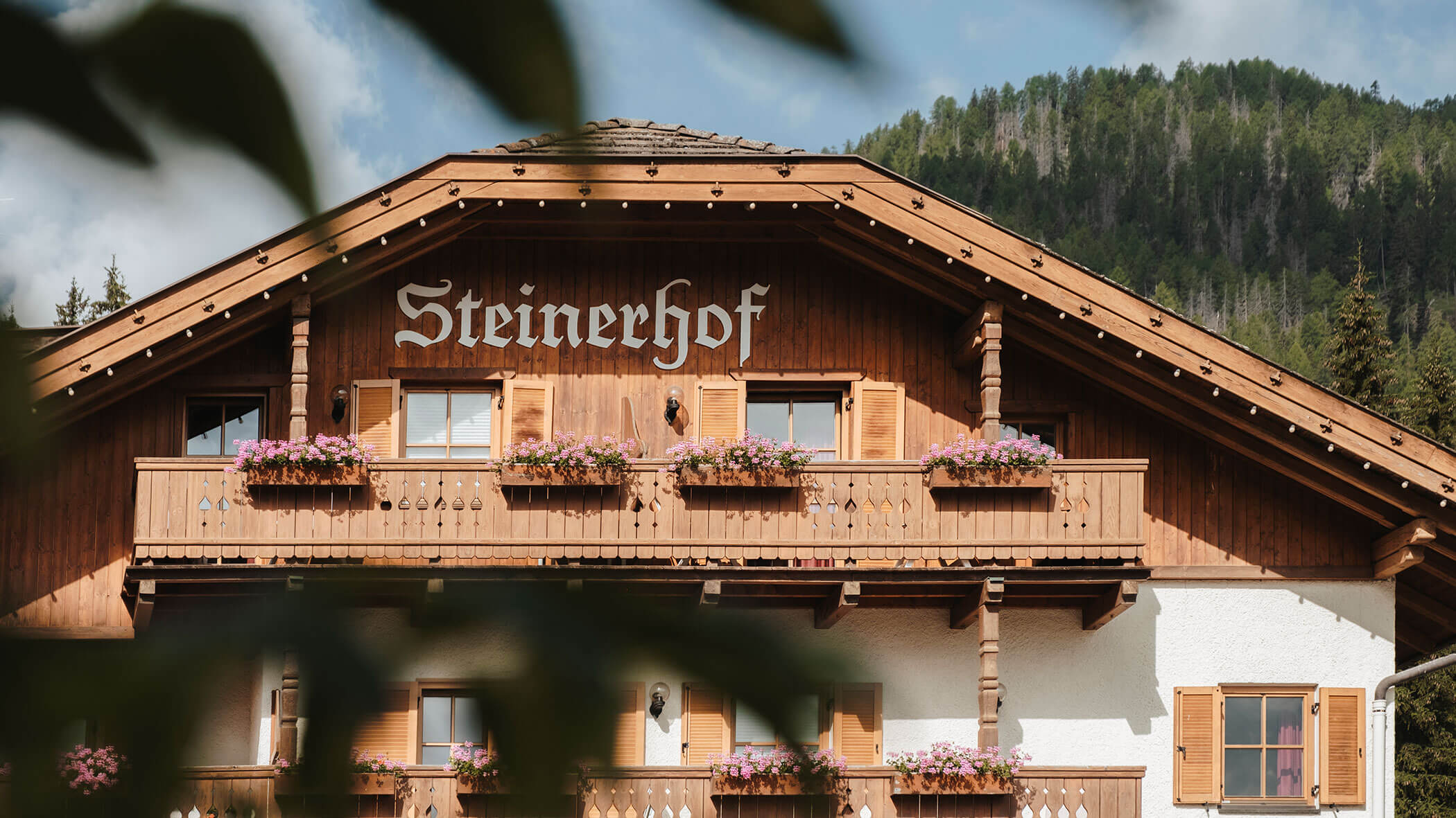 Holzchalet mit Balkonen, Blumenkästen und einem „Steinerhof“-Schild vor dem Hintergrund eines bewaldeten Berges. - Hotel Steinerhof