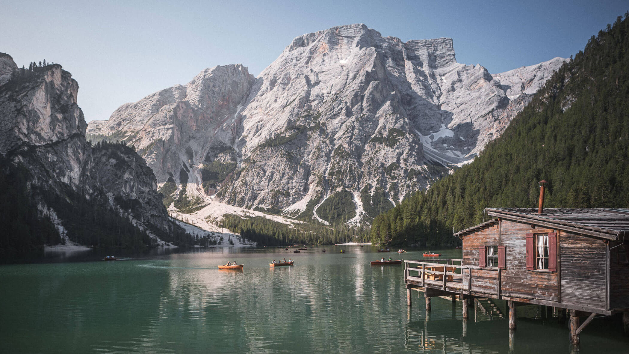 Eine Holzhütte an einem See, mit Booten und hohen, felsigen Bergen im Hintergrund unter einem klaren Himmel. - Hotel Steinerhof