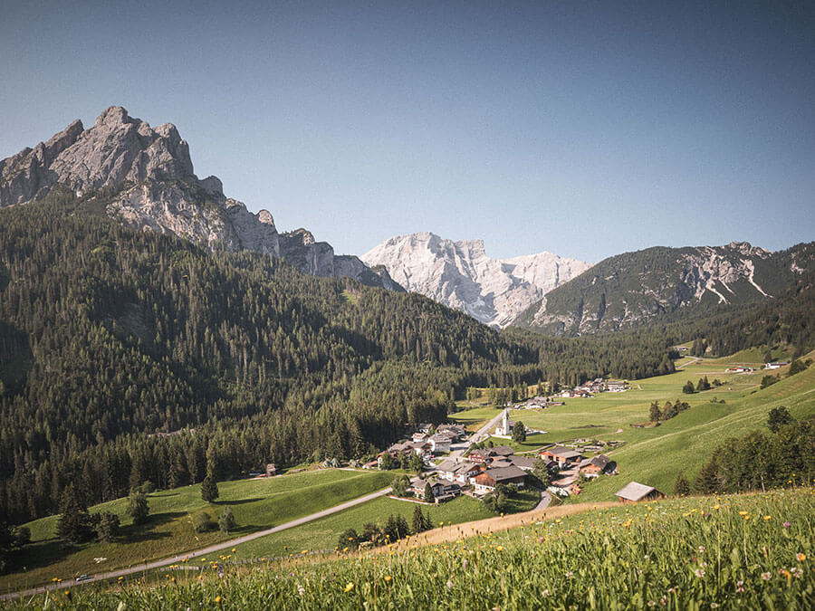 Ein Dorf mit vereinzelten Häusern liegt in einem grünen Tal, umgeben von Bergen und Wäldern, unter einem klaren Himmel. - Hotel Steinerhof