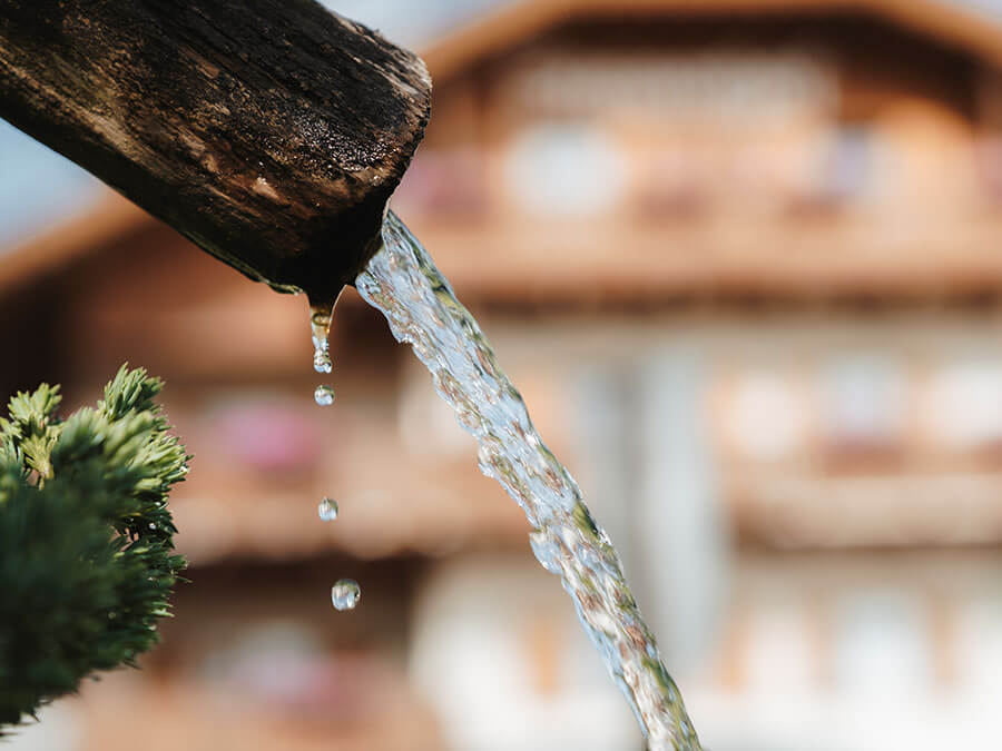 Aus einem hölzernen Auslauf fließt klares Wasser, im Hintergrund ist ein verschwommenes, rustikales Gebäude zu sehen. - Hotel Steinerhof