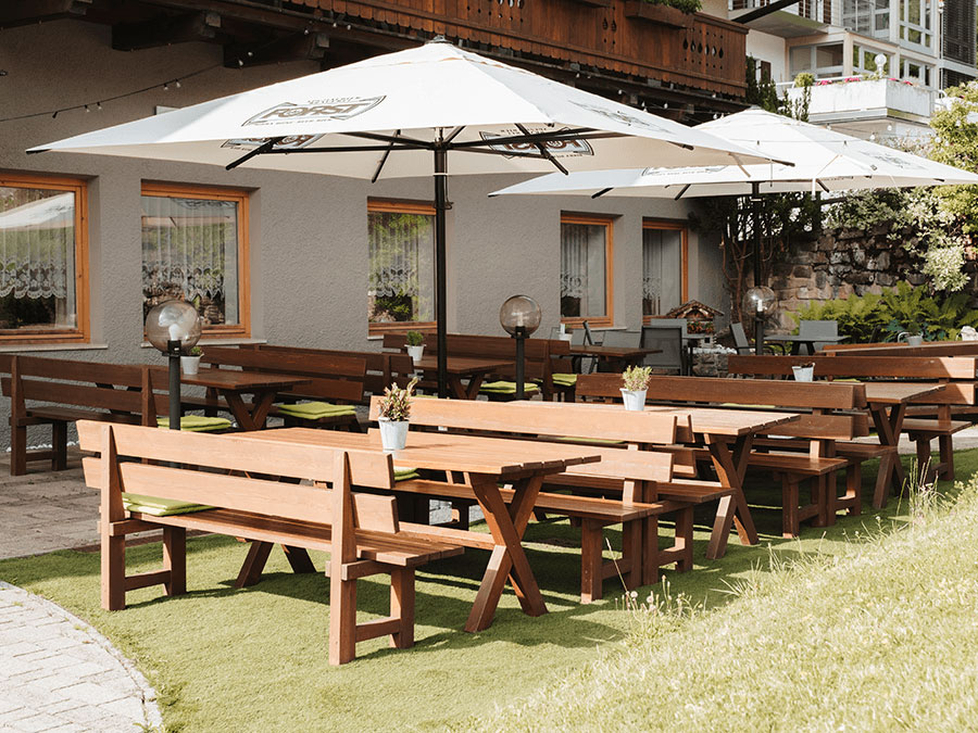 Café im Freien mit Holzbänken, Tischen, Sonnenschirmen, Topfpflanzen und einem Gebäude im Hintergrund. - Hotel Steinerhof