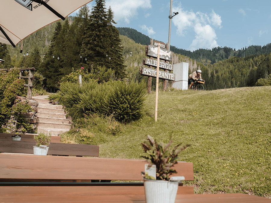Holzbänke, Topfpflanzen und ein Schild mit der Aufschrift „ALBERGO RISTORANTE“ in einer malerischen, hügeligen Umgebung im Freien. - Hotel Steinerhof