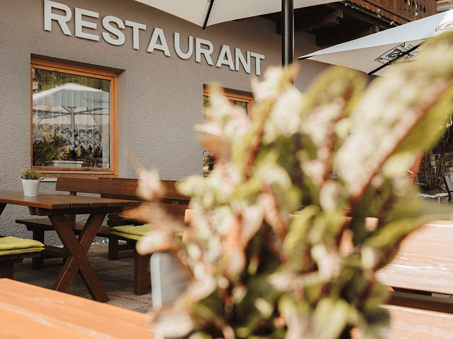 Terrasse eines Restaurants im Freien mit Holztischen, Bänken, Sonnenschirmen und einer unscharfen Pflanze im Vordergrund. - Hotel Steinerhof